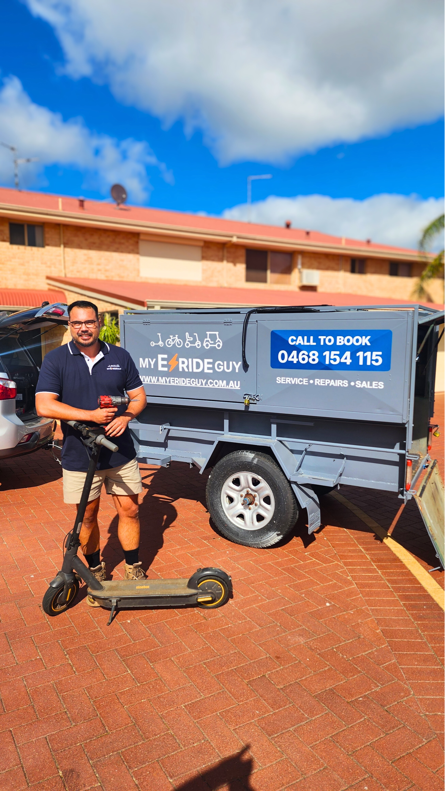 Man standing next to a mobile repair trailer for My ERide Guy, holding an e-scooter, with contact information for scooter repairs visible on the trailer.