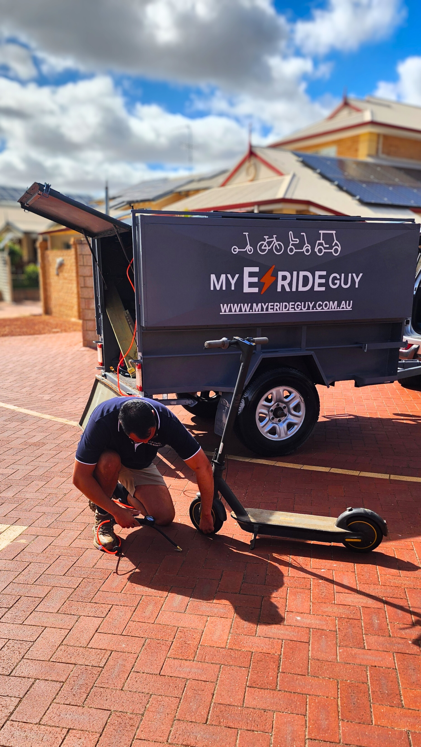 Person repairing an electric scooter next to a branded service trailer for My E Ride Guy, emphasizing mobile e-scooter repair services in a sunny outdoor setting.